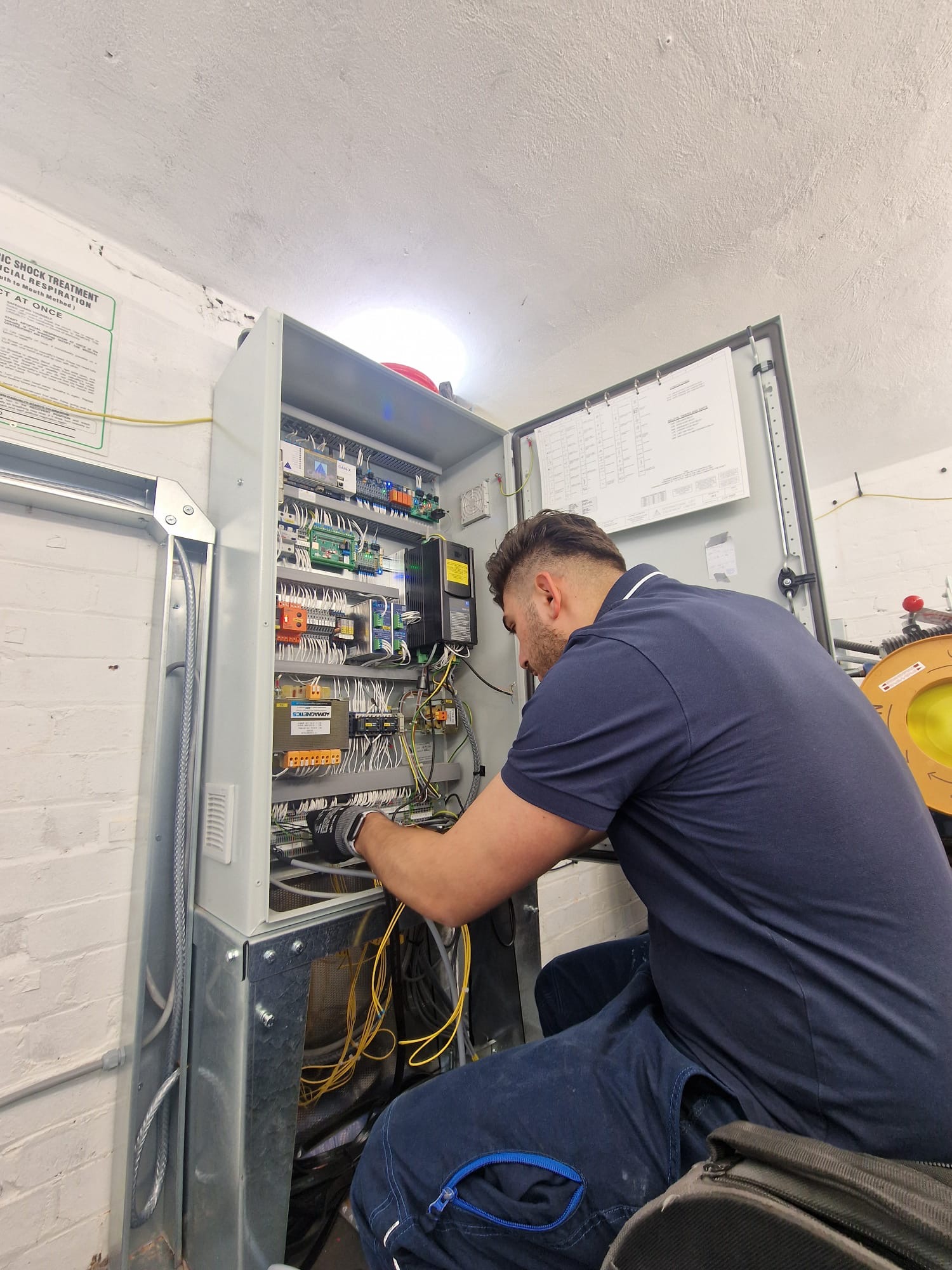 A man wearing a navy blue polo and gloves is working on the wiring inside an open gray electrical control panel mounted on a white brick wall, with various colored wires, circuit boards, and components visible.