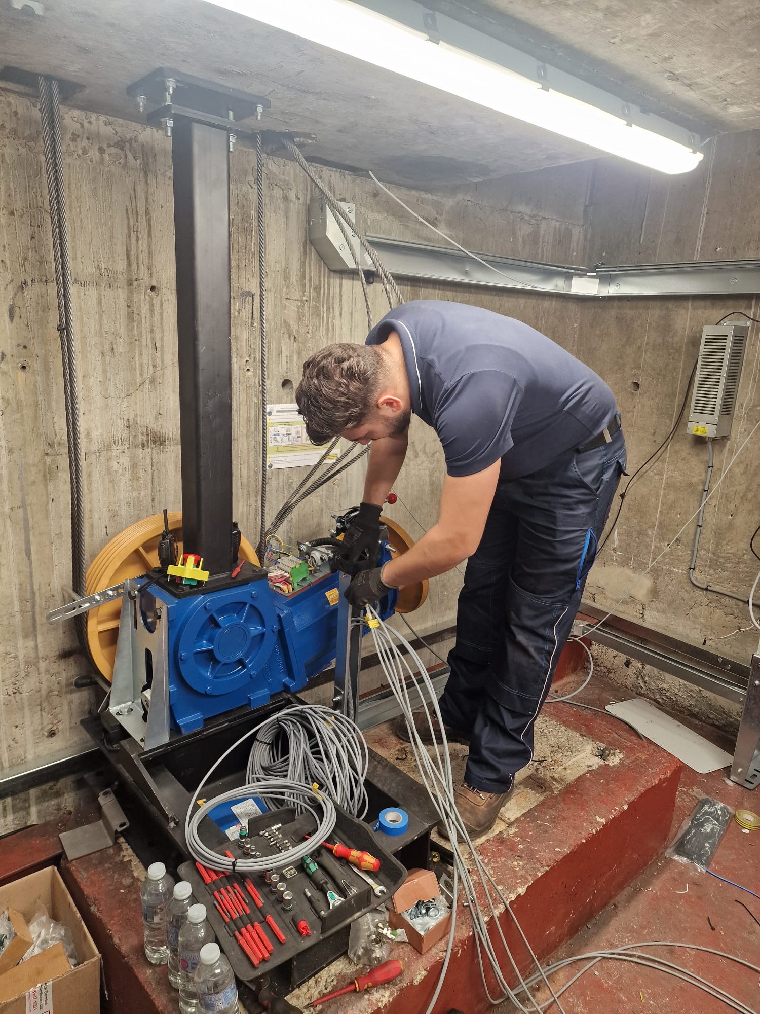 Technician in a concrete room working on a blue industrial machine with large yellow pulleys, handling several gray cables, surrounded by tools and water bottles.