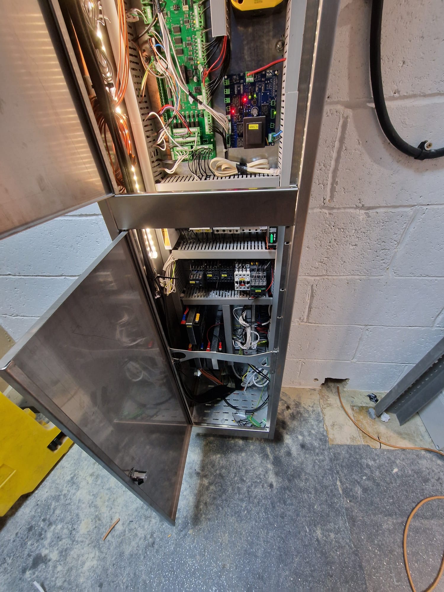 Open metal electrical control panel mounted on a wall showing multiple circuit boards, wiring, relays, and electronic components inside with cables running from the panel and a concrete wall background.