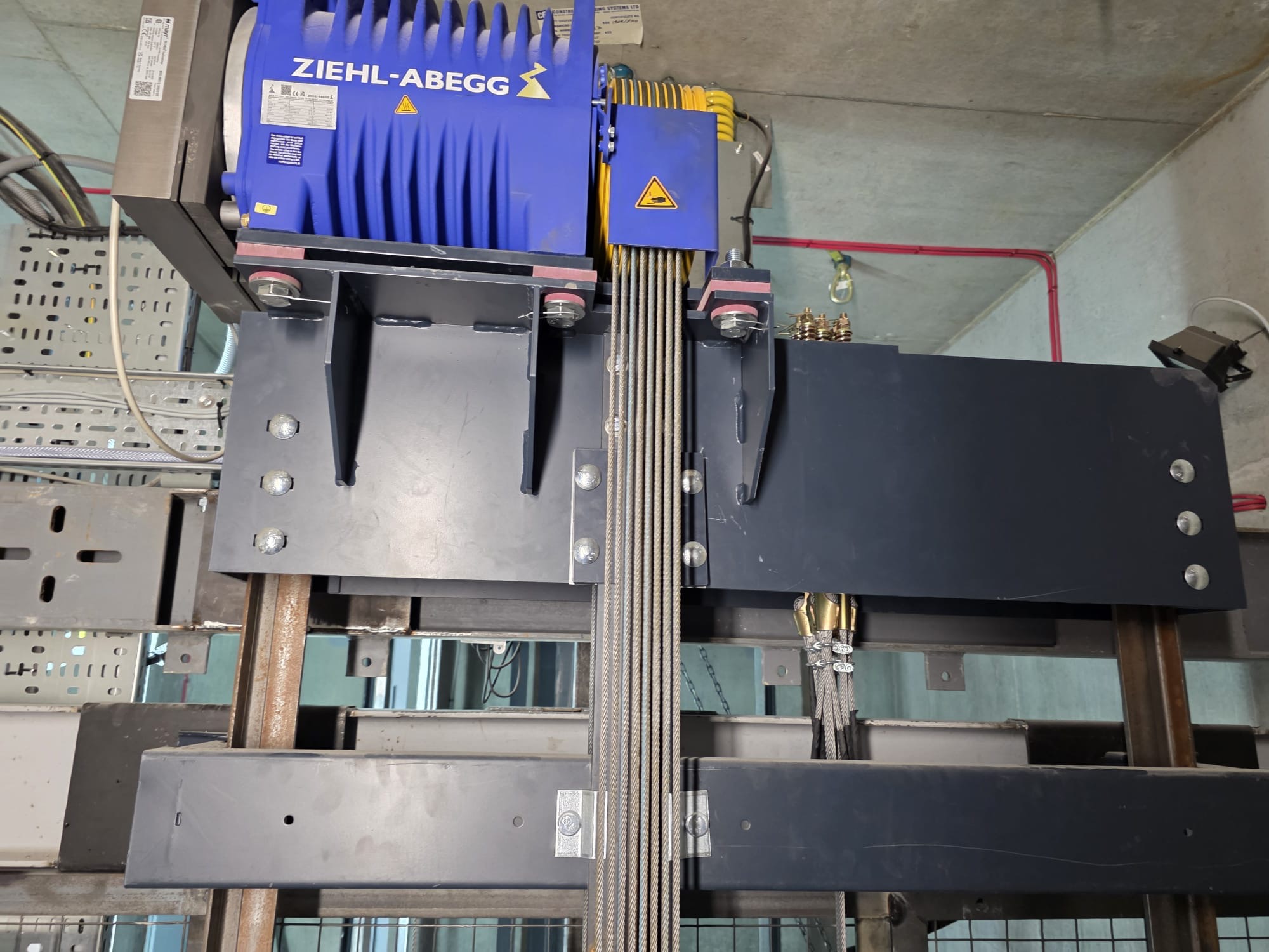 Close-up of a Ziehl-Abegg branded blue motor with multiple thick steel cables wrapped around pulleys, mounted on a metal frame inside an industrial or elevator shaft setting.