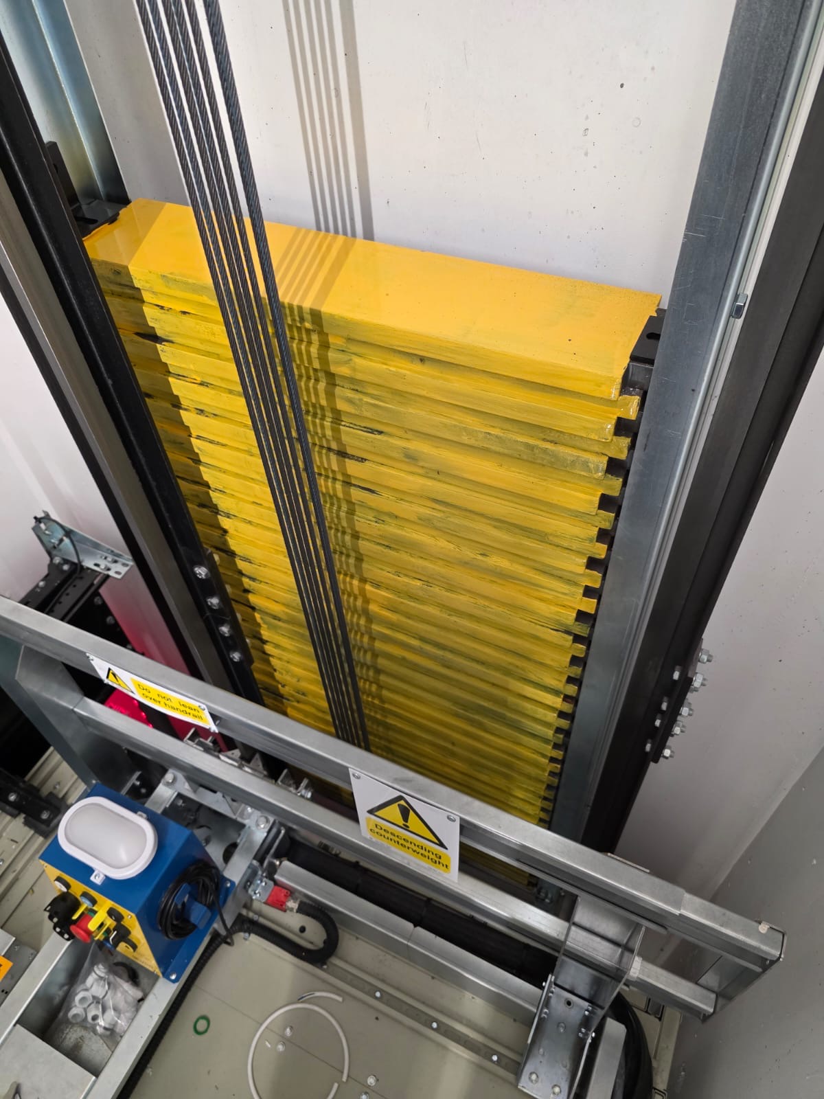 Close-up of inside an elevator shaft showing yellow elevator counterweights connected by multiple steel cables and metal framework with warning signs.