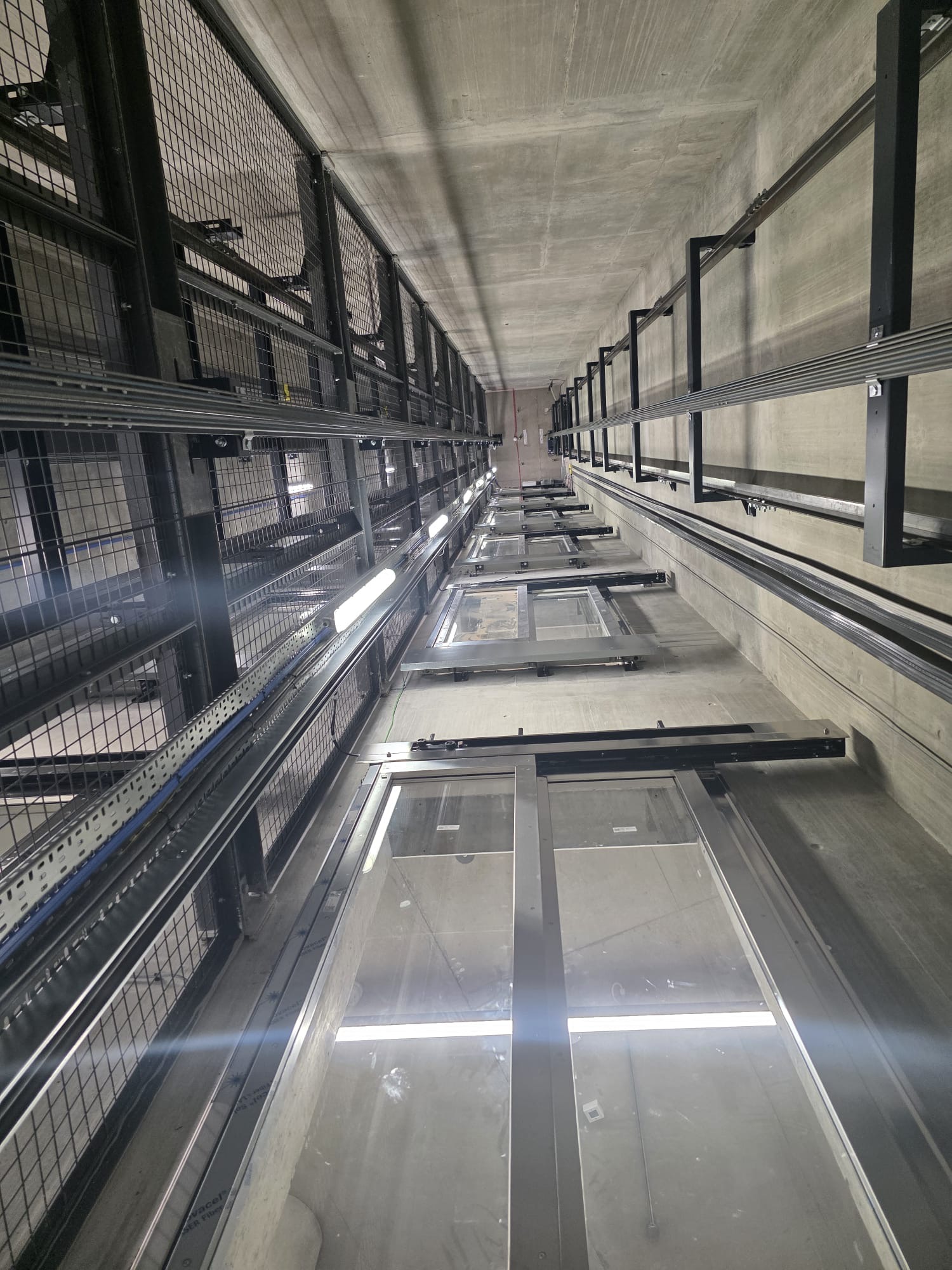 View looking up an elevator shaft showing metal rails, cables, and the framework for elevator doors fixed to concrete walls.