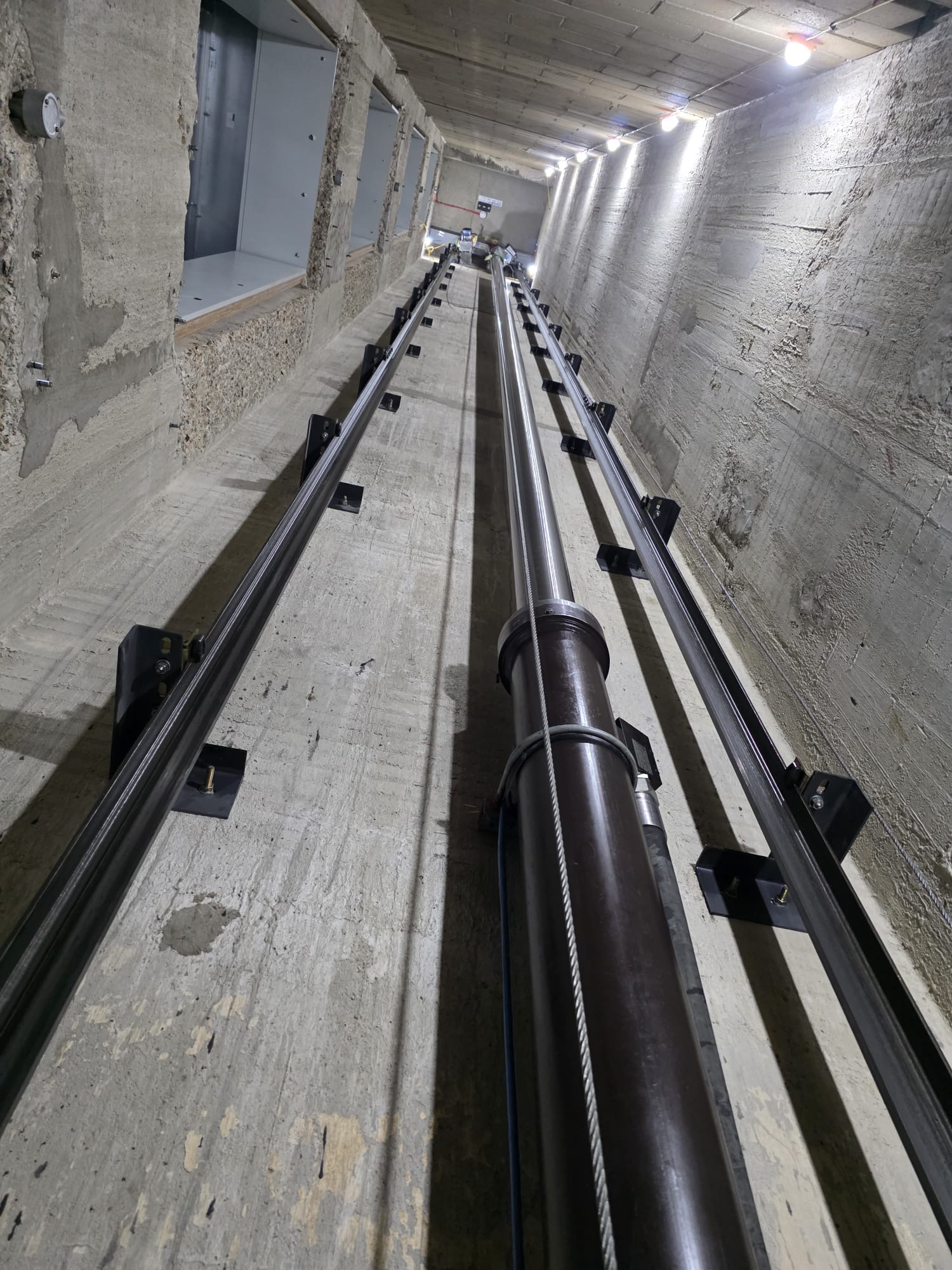View looking up inside a tall, narrow elevator shaft with concrete walls, metal rails, and a central cylindrical shaft component supported by cables.