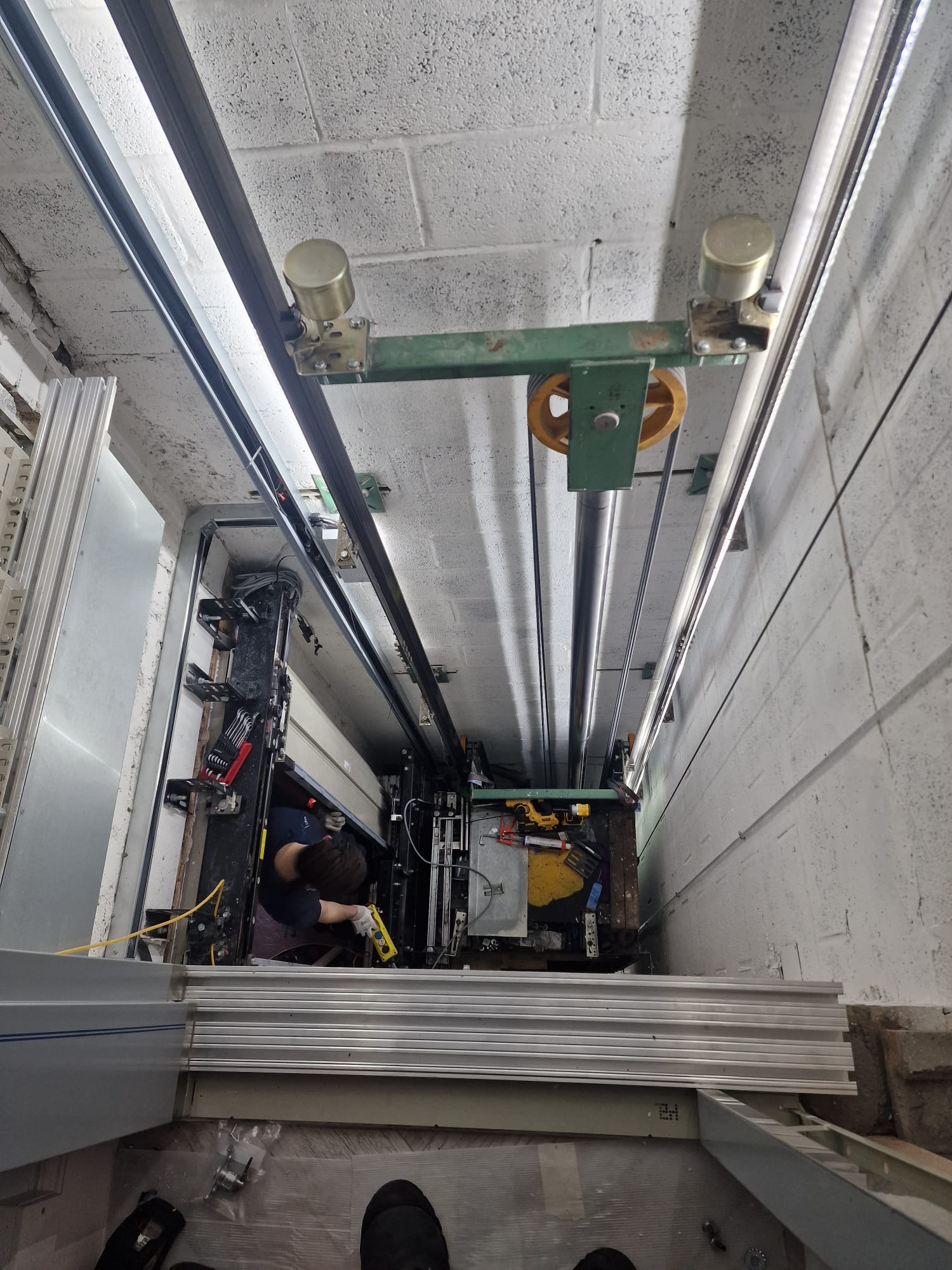 Technician wearing gloves working inside an elevator shaft, handling tools and equipment, viewed from above.