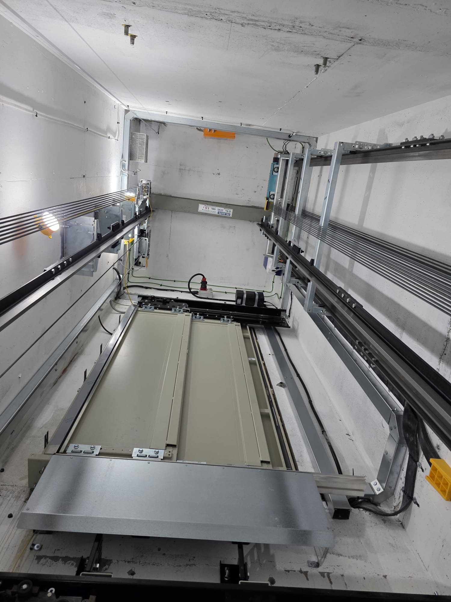 Interior view of an elevator shaft showing elevator doors, metal rails, cables, and mechanical components mounted on white concrete walls and ceiling.
