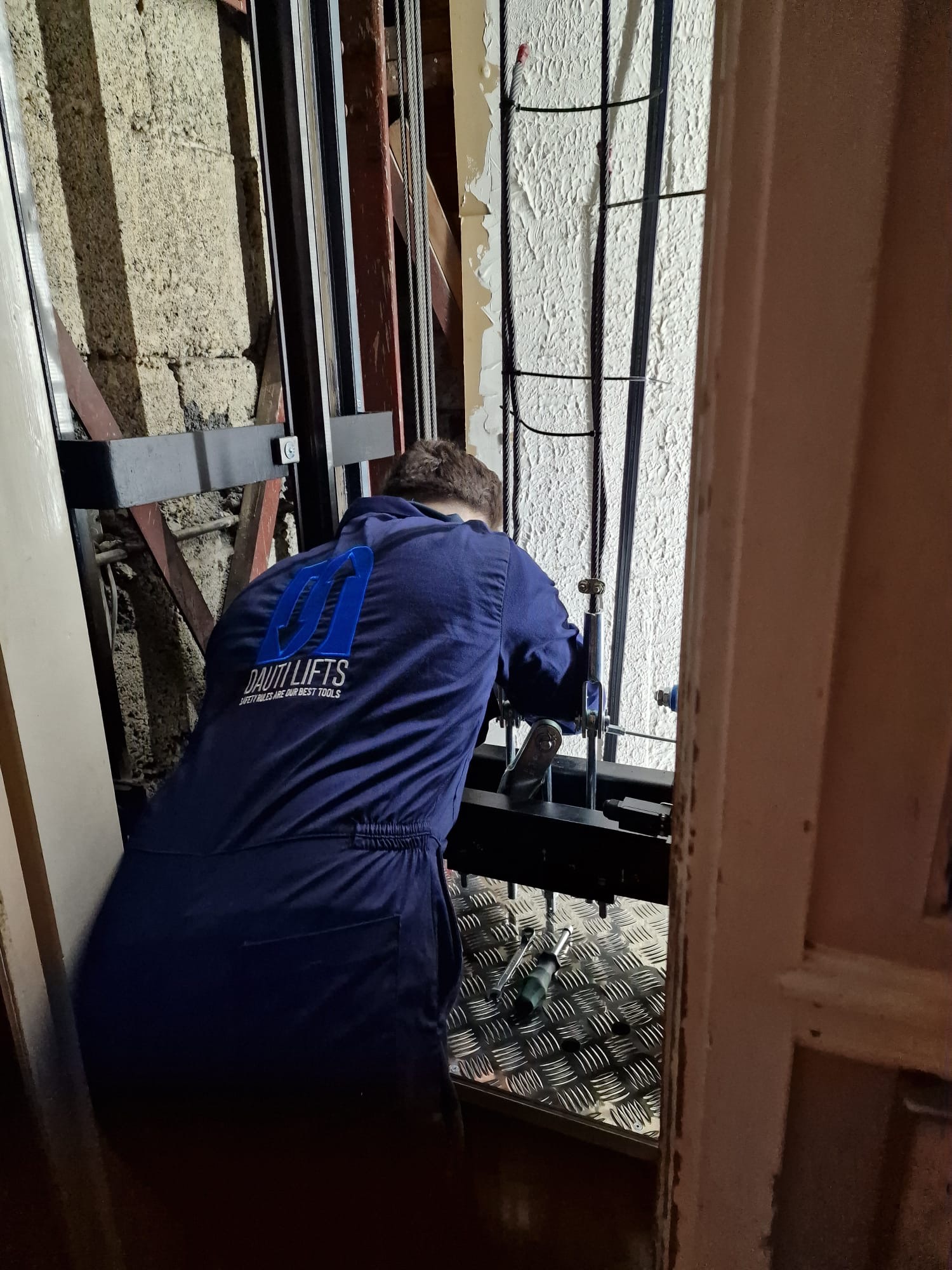 Technician wearing a dark blue jumpsuit with the logo 'Dauti Lifts' working on elevator machinery inside an elevator shaft.