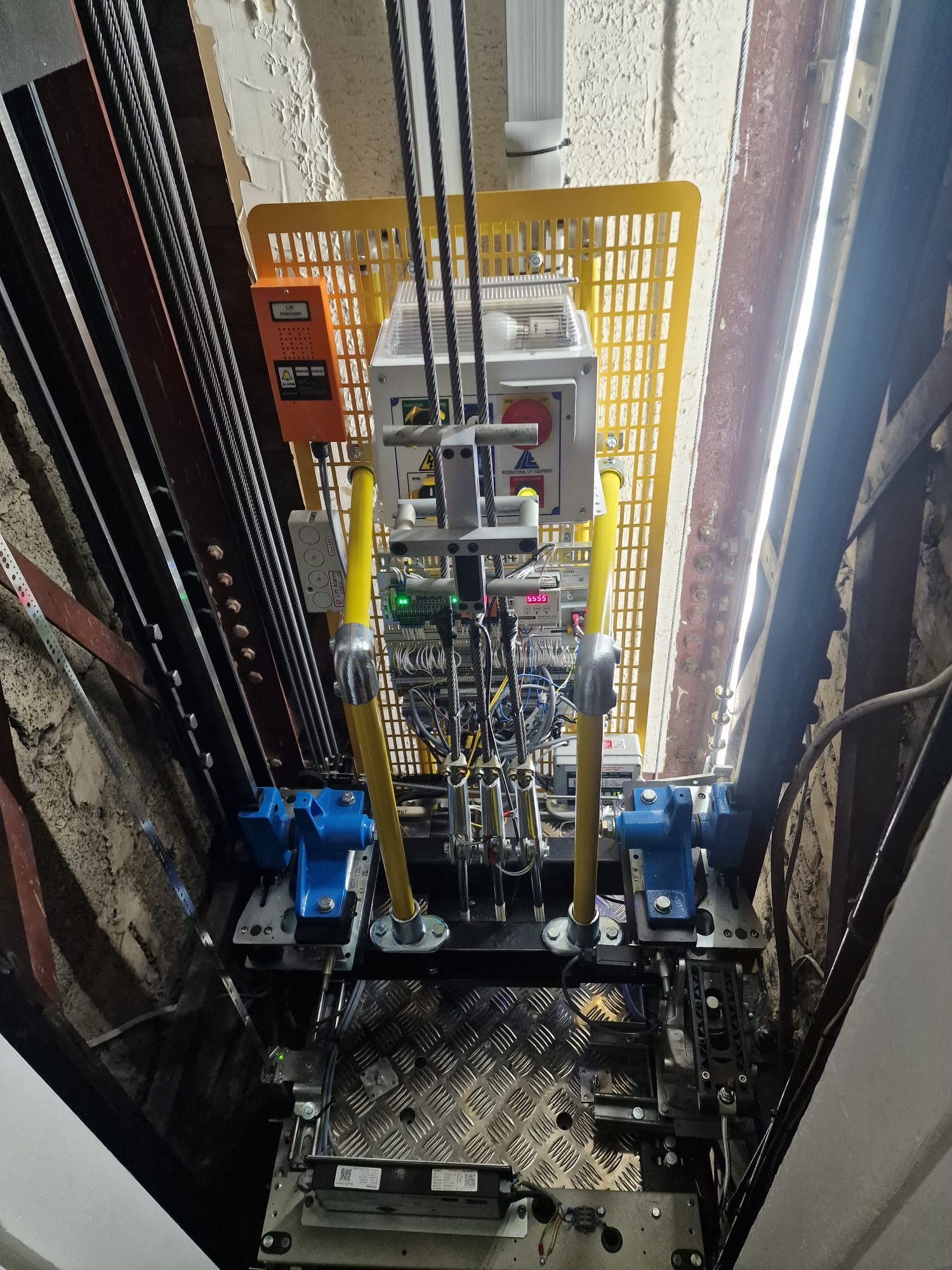 View inside an elevator shaft showing complex mechanical and electrical components, including multiple steel cables, yellow safety bars, blue mounted parts on either side, control panels with wiring, and a grated metal platform.