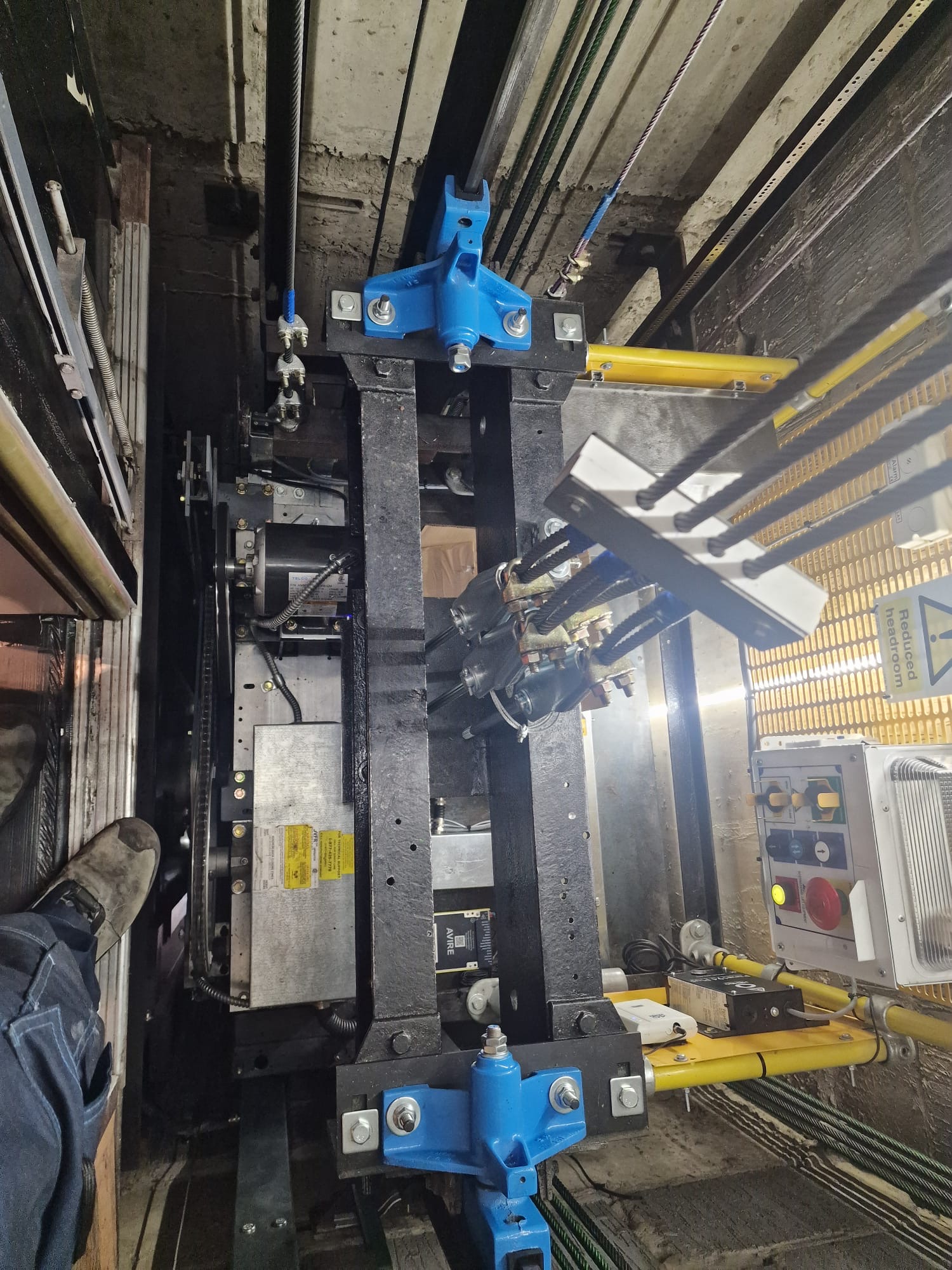 Interior view of an elevator shaft showing mechanical components including blue mounting brackets, black metal beams, cables, pulleys, a control panel with buttons, and a foot wearing a work boot on the left side.