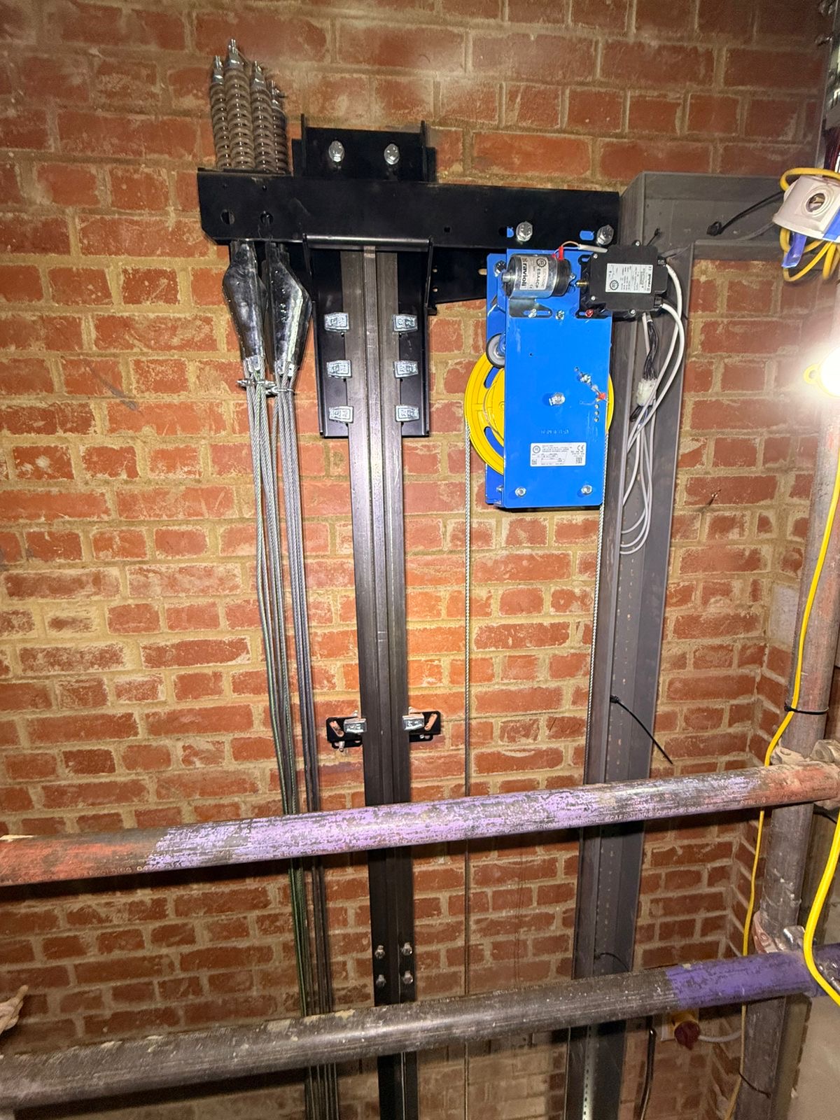 Mechanical assembly mounted on a brick wall featuring a blue metal box with a yellow spool and various electrical components, alongside black vertical metal rails and steel cables, with two horizontal pipes in the foreground.
