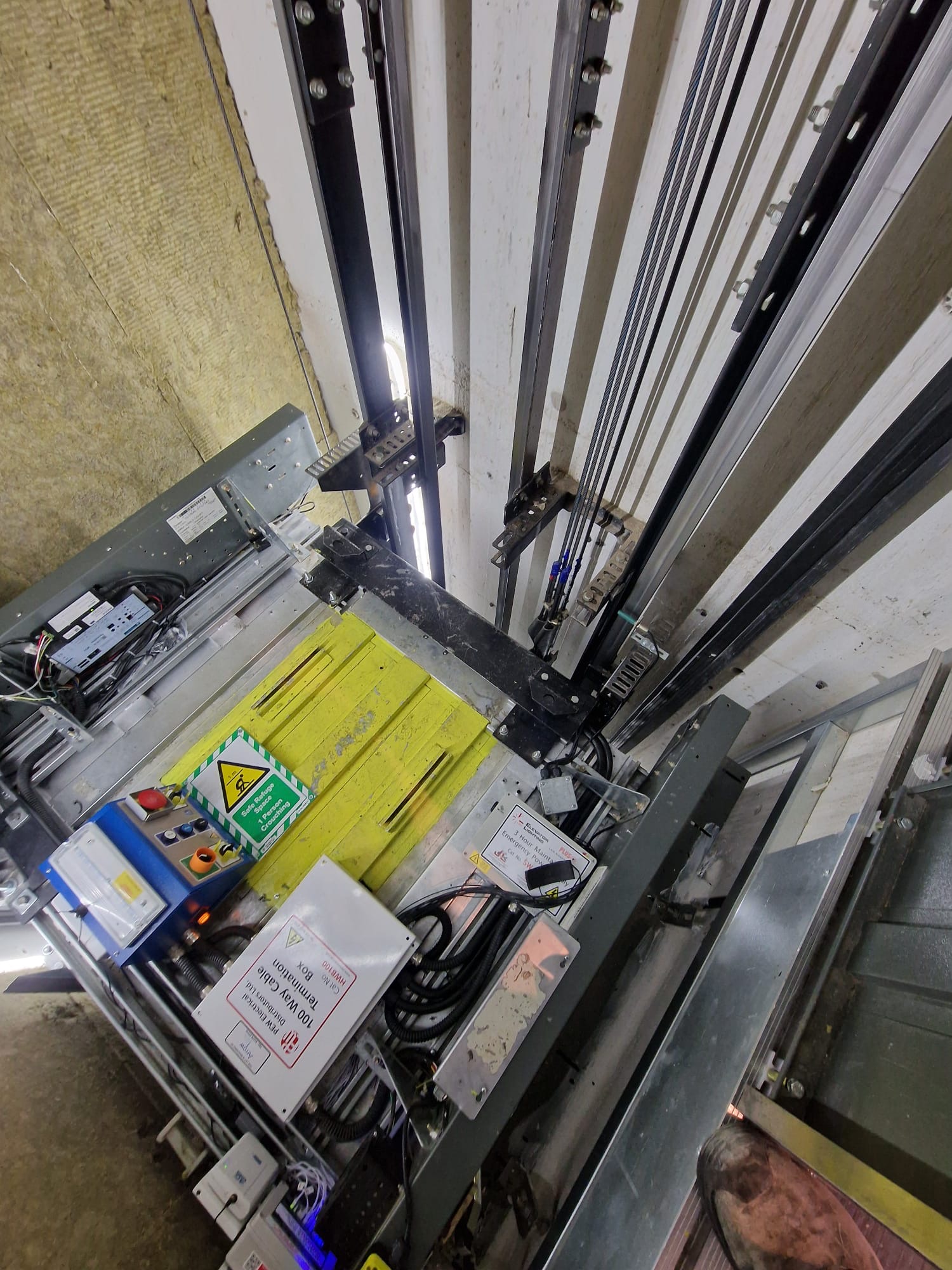 View inside an elevator shaft showing elevator platform with control boxes, cables, rails, and metal structures against a concrete wall.