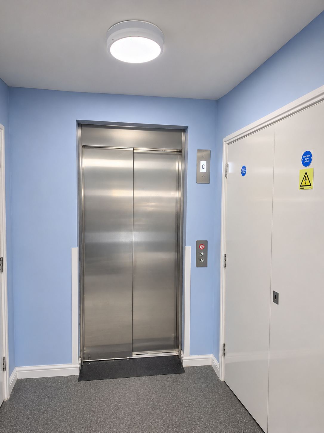 Indoor elevator with closed stainless steel doors set in a light blue wall, with a control panel showing buttons and a floor indicator lit on 'G', next to a white door with safety warning stickers.