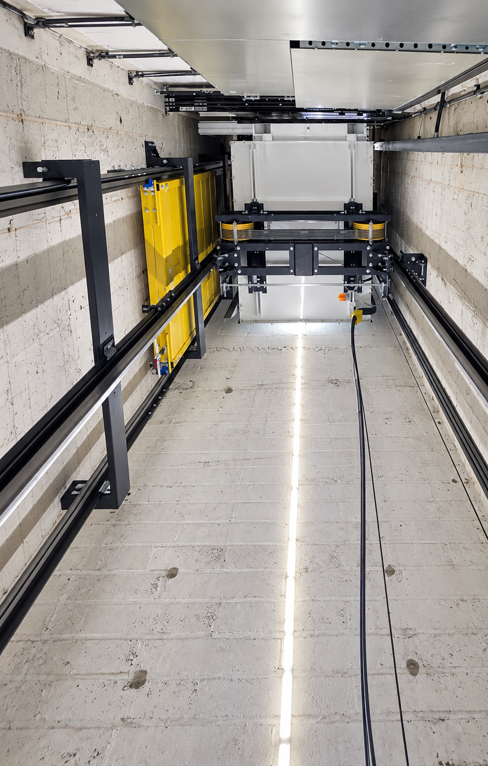 Inside view of a modern elevator shaft showing mechanical components, metal tracks, a yellow platform, and a vertical strip light on the concrete wall.
