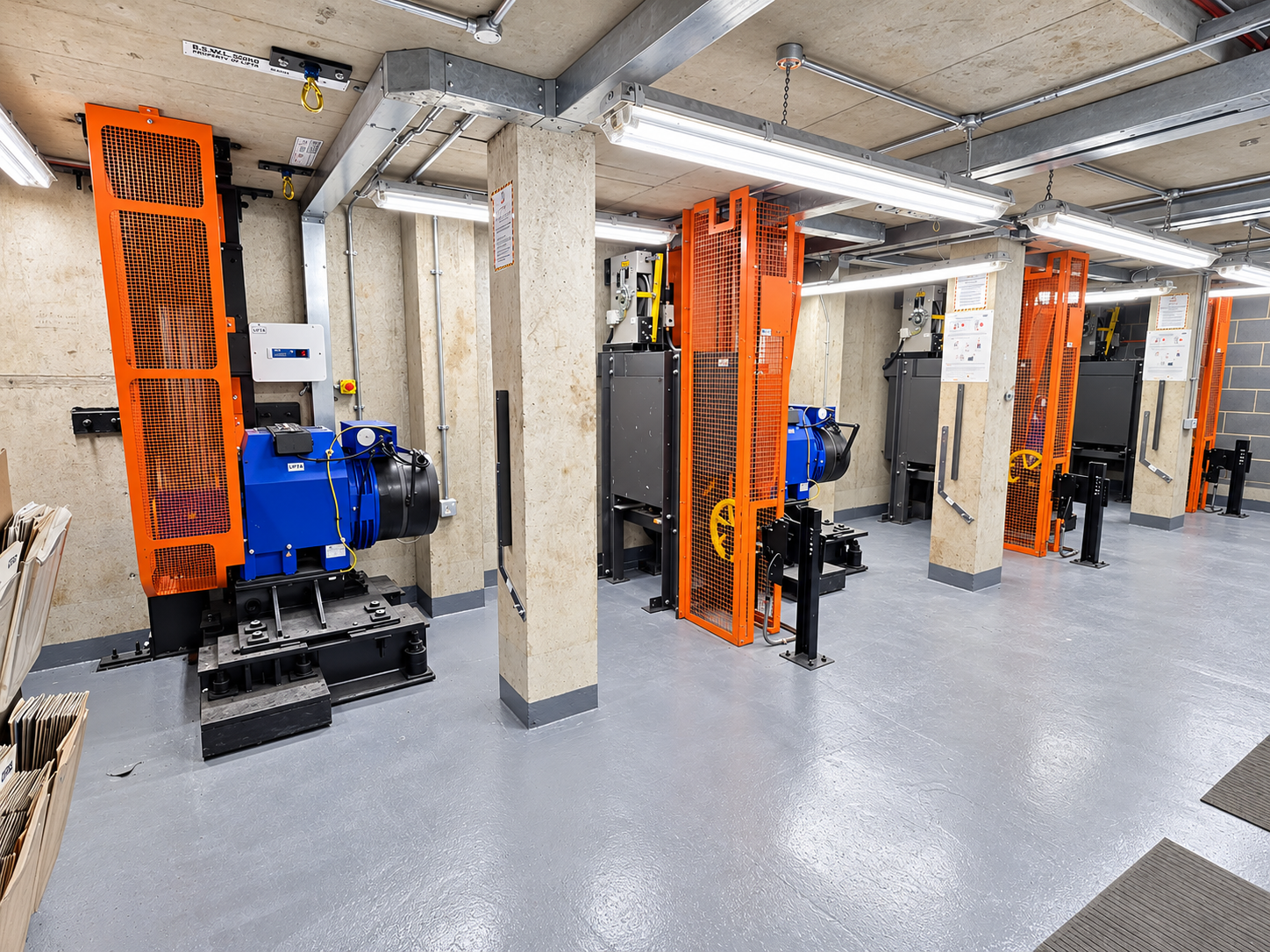 Industrial room with multiple mechanical lift systems featuring blue motors and orange protective cages, mounted on a gray polished floor with beige walls and concrete pillars.