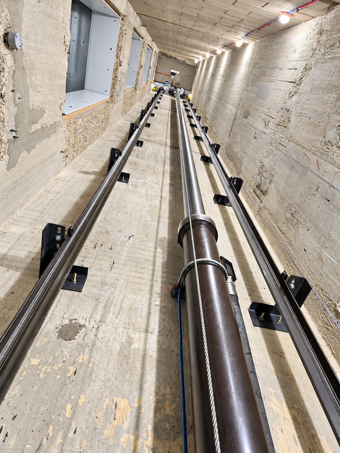 Interior view of an elevator shaft showing two vertical metal rails mounted on the concrete walls, a thick central metal cylinder with a steel cable running alongside it, and rough concrete walls with electrical wiring and light bulbs lining the ceiling.