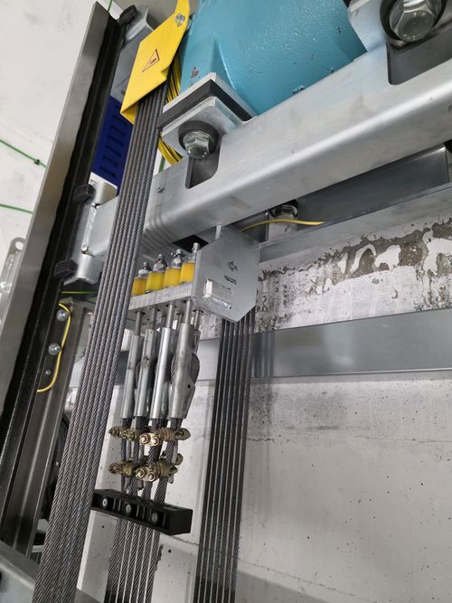 Close-up of elevator steel cables secured by clamps and connected to a metal support frame mounted on a wall with visible concrete and peeling paint.