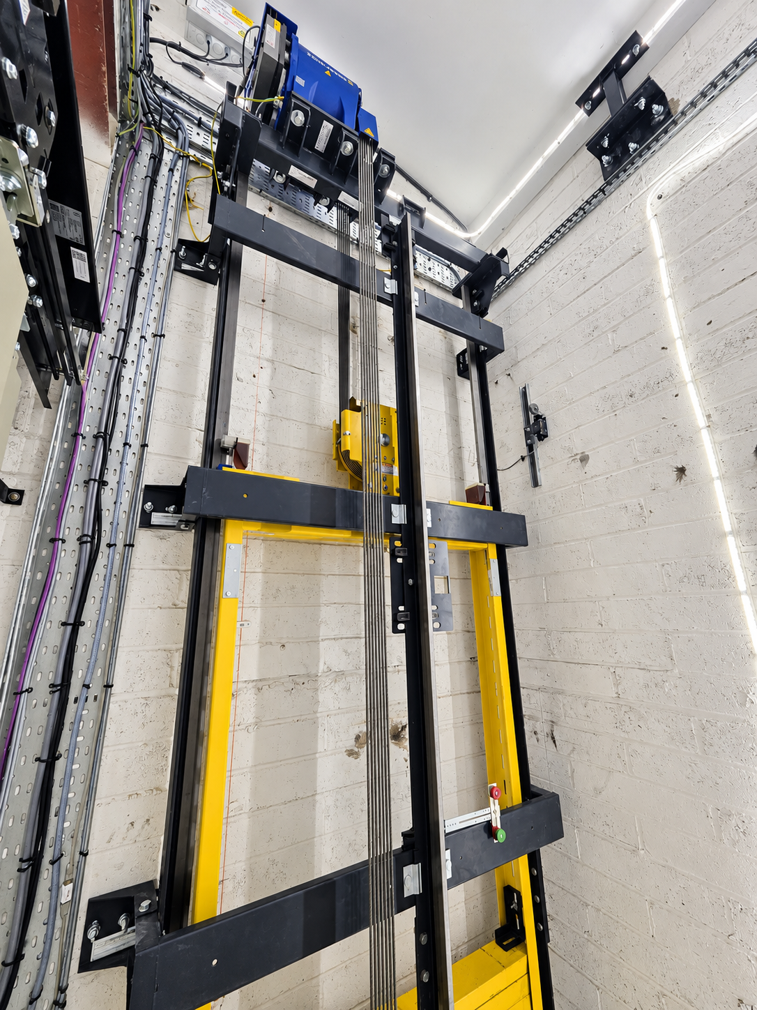 Interior of an elevator shaft showing the mechanical components including steel cables, black and yellow metal framework, and electrical wiring mounted on white brick walls.