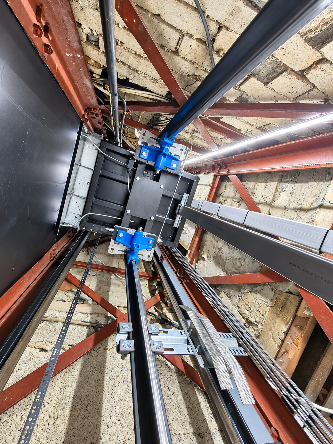 Inside view of an elevator shaft showing metal rails, blue mechanical parts, cables, and a concrete ceiling with red steel support beams.