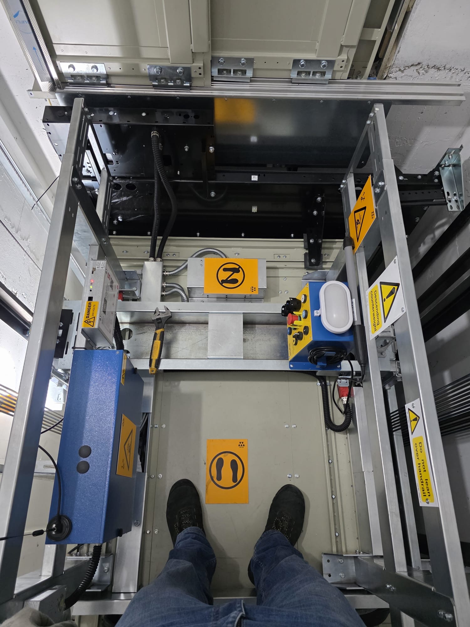 View looking down at a person standing inside a metal-framed elevator shaft or industrial lift platform with control panels, warning signs, a wrench, and a yellow floor marker for foot placement.