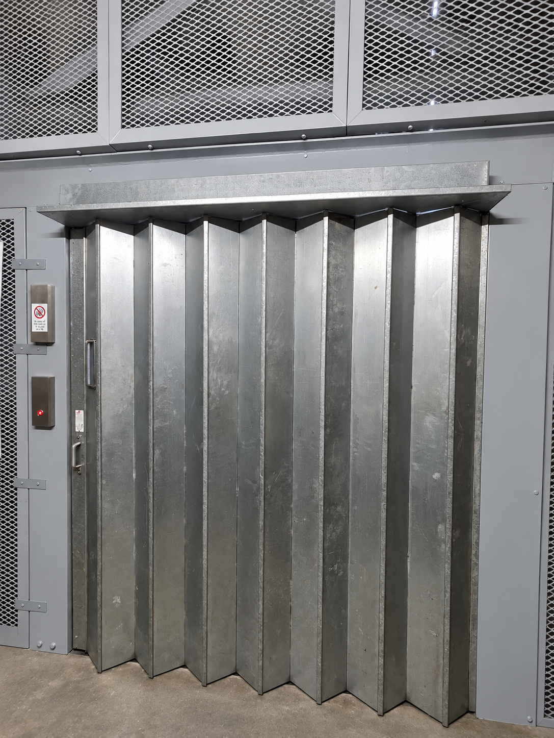 Metal accordion-style folding security door in a gray industrial setting with a handle on the left side and a red indicator light on a nearby panel.