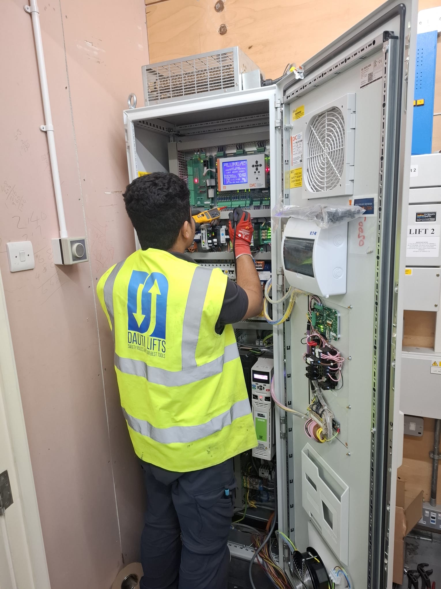 Technician wearing a bright yellow high-visibility vest with blue logo and dark pants working inside an open electrical control panel with circuit boards and wiring in a utility room.