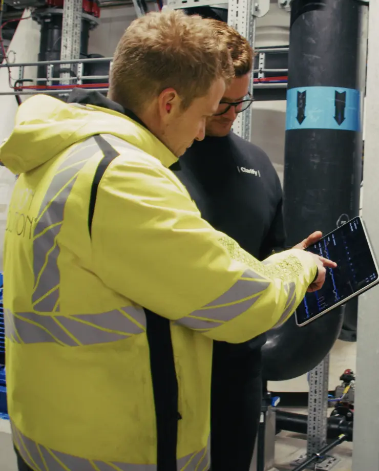 Two men in a mechanical room examining data on a tablet; one wearing a yellow reflective jacket and the other in a dark shirt with glasses.