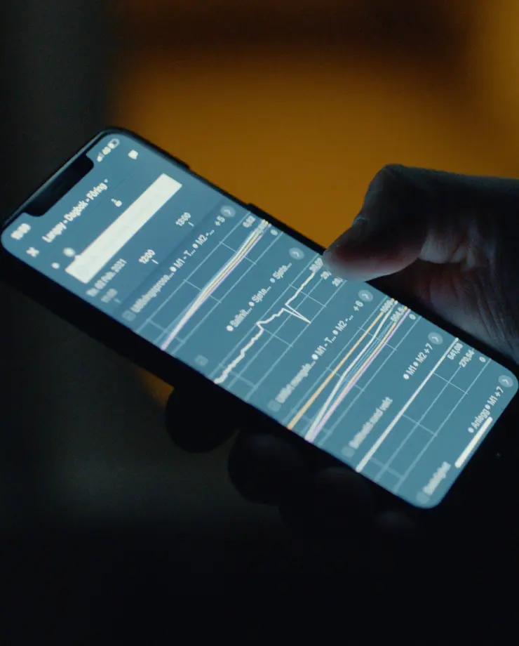 Hand holding a smartphone displaying multiple line graphs and charts with data points in a dark environment.