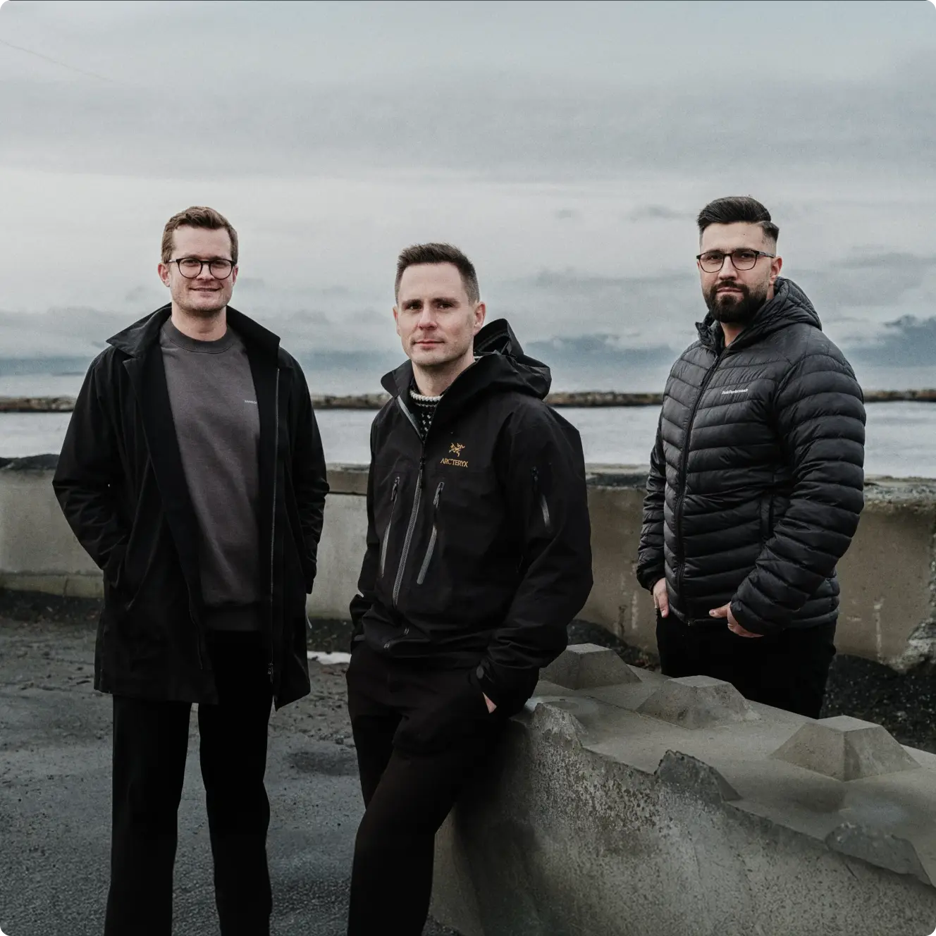 Three men wearing black jackets standing outdoors near a concrete barrier with a cloudy sky and body of water in the background.