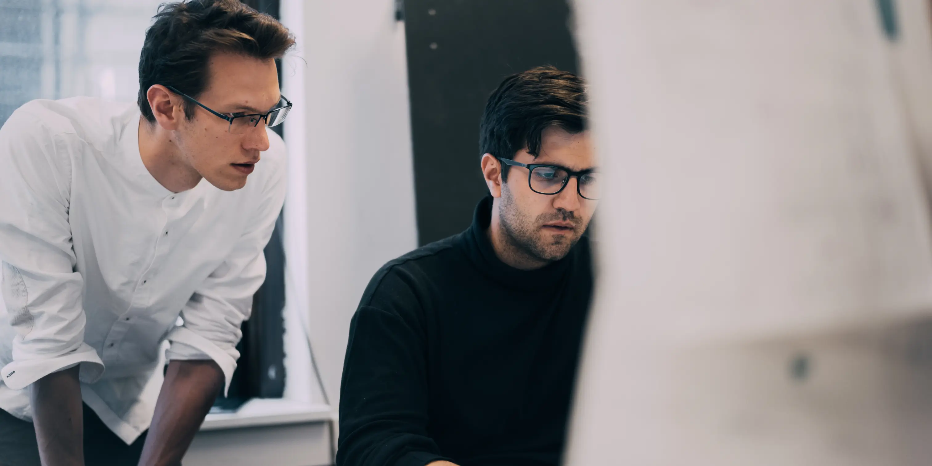 Two men wearing glasses working together at a desk, one seated and focused on a computer while the other leans forward, observing.