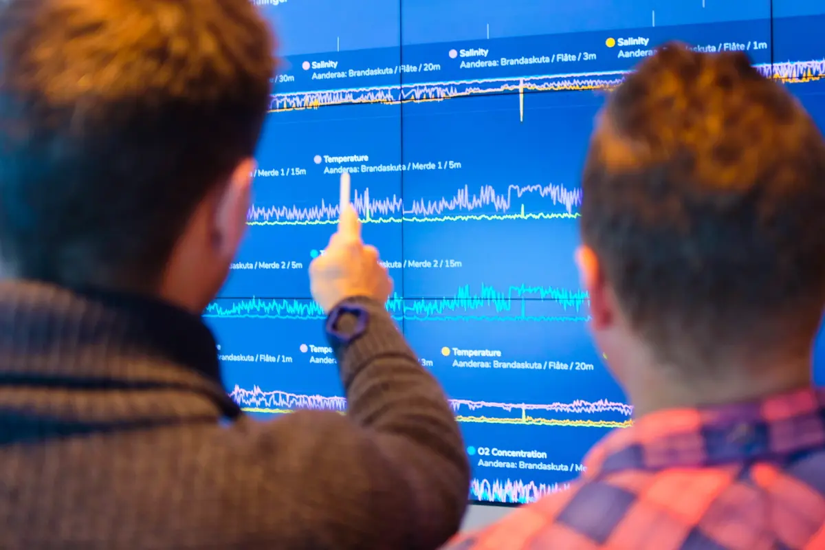 Two people viewed from behind observe and point at a large screen displaying multiple data graphs with labeled lines indicating salinity, temperature, and O2 concentration over different depths.