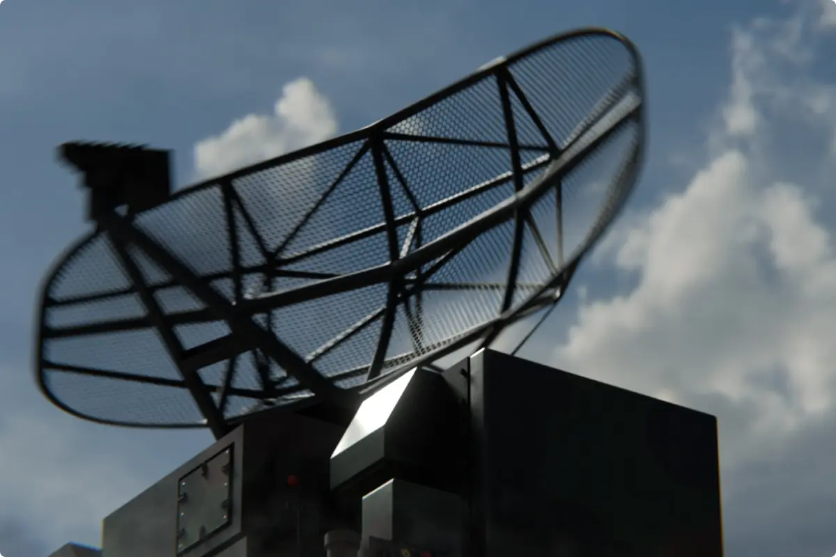 Close-up of a black radar dish with a mesh reflector mounted on a large dark base against a partly cloudy sky.