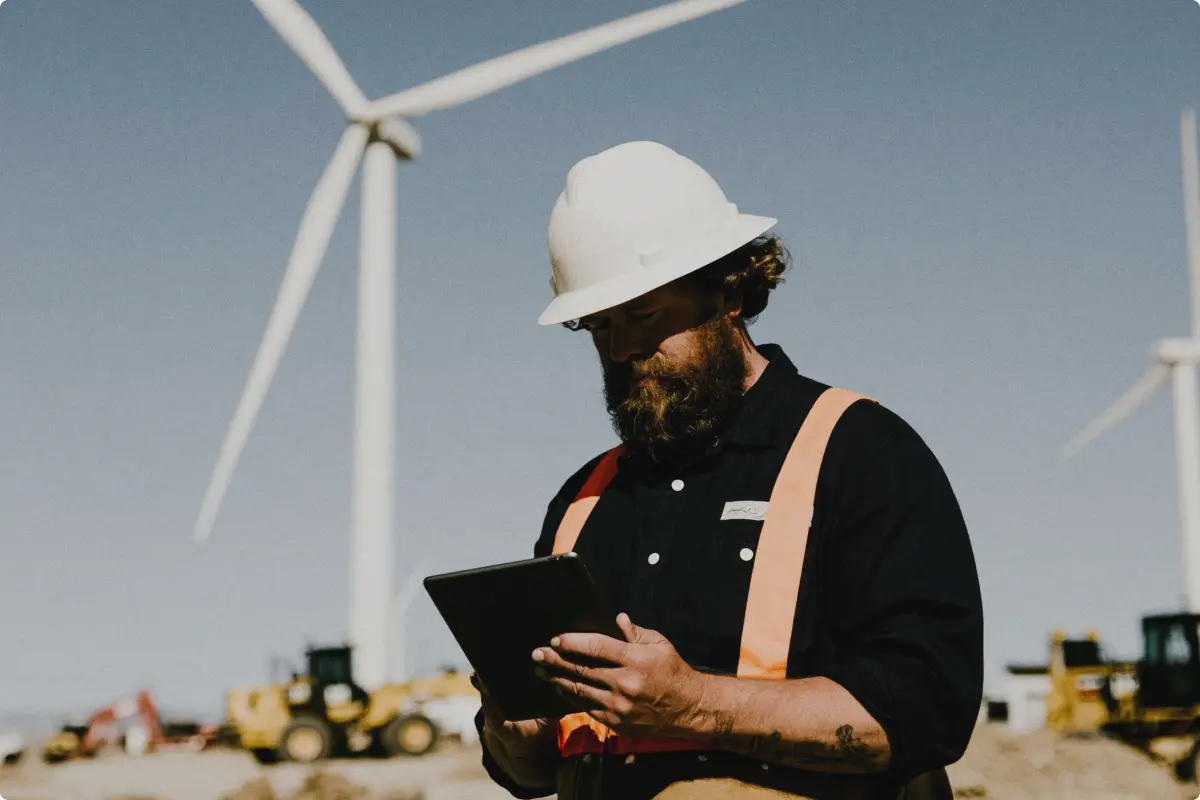 A bearded construction worker wearing a white hard hat and orange safety suspenders uses a tablet at a wind farm with turbines and construction equipment in the background.