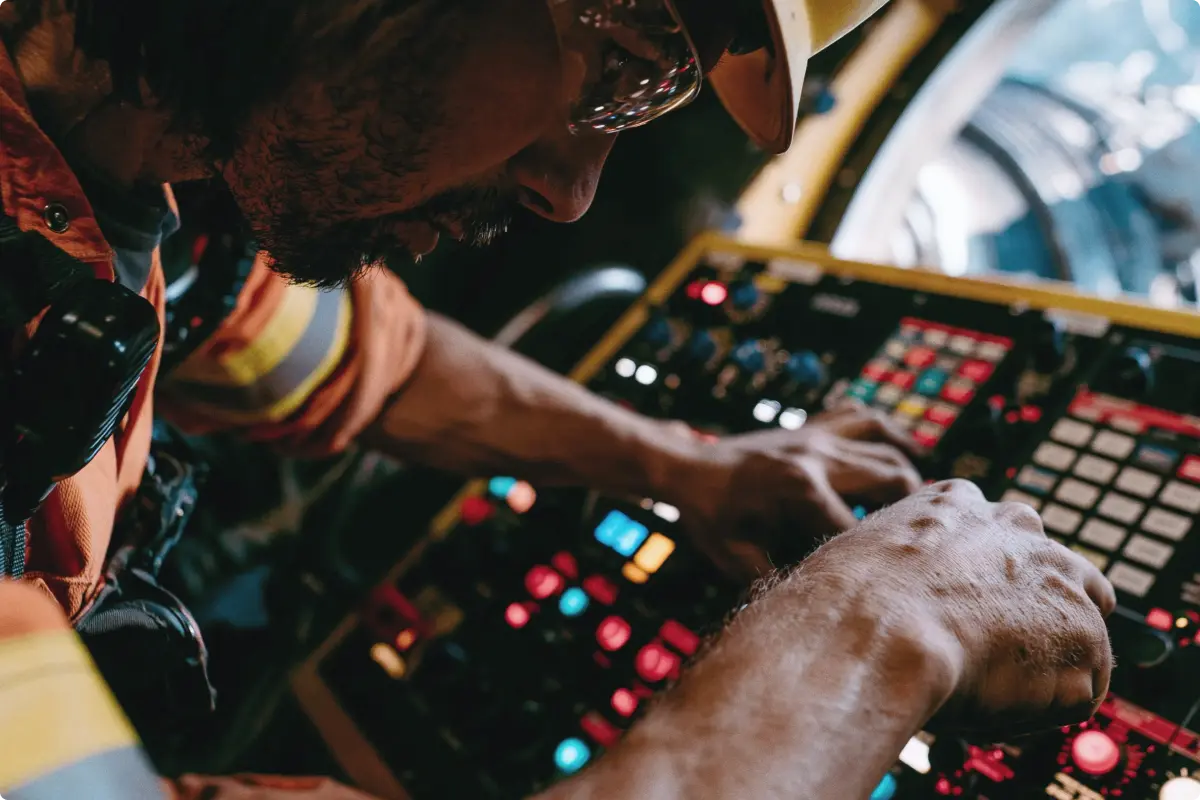 Man in safety helmet and glasses operating control panel with numerous buttons and knobs, focused on adjusting settings.