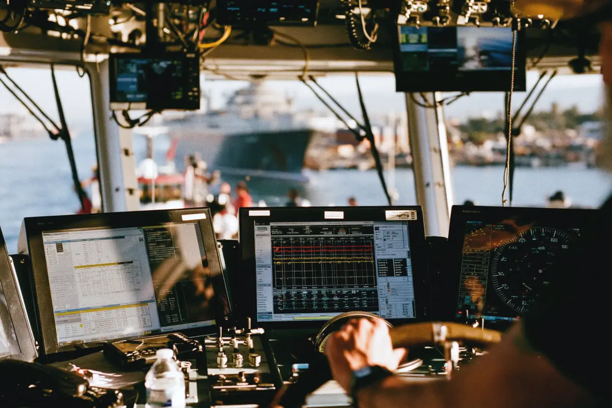 View from inside a ship's bridge showing a person operating the ship's controls with multiple digital navigation and monitoring screens, and an out-of-focus large vessel visible through the windows in the background.