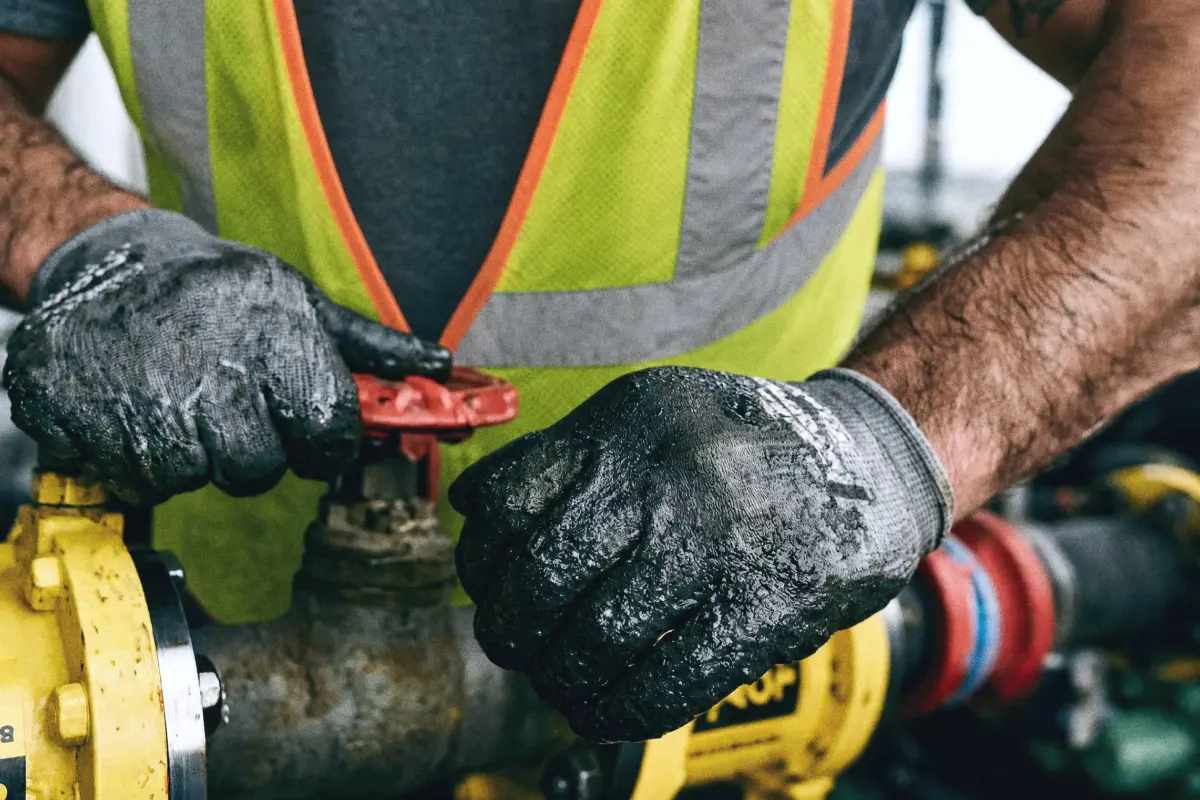 Close-up of a worker in a reflective safety vest and dirty gloves operating a red valve on yellow industrial piping.