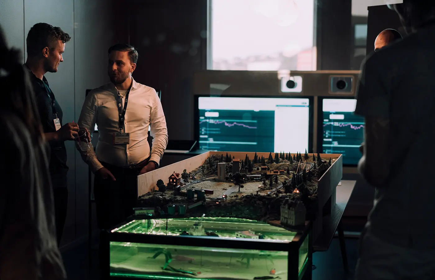 Two men wearing badges are conversing near a detailed miniature landscape model with terrain, trees, and buildings, in a dimly lit room with computer screens displaying graphs in the background.