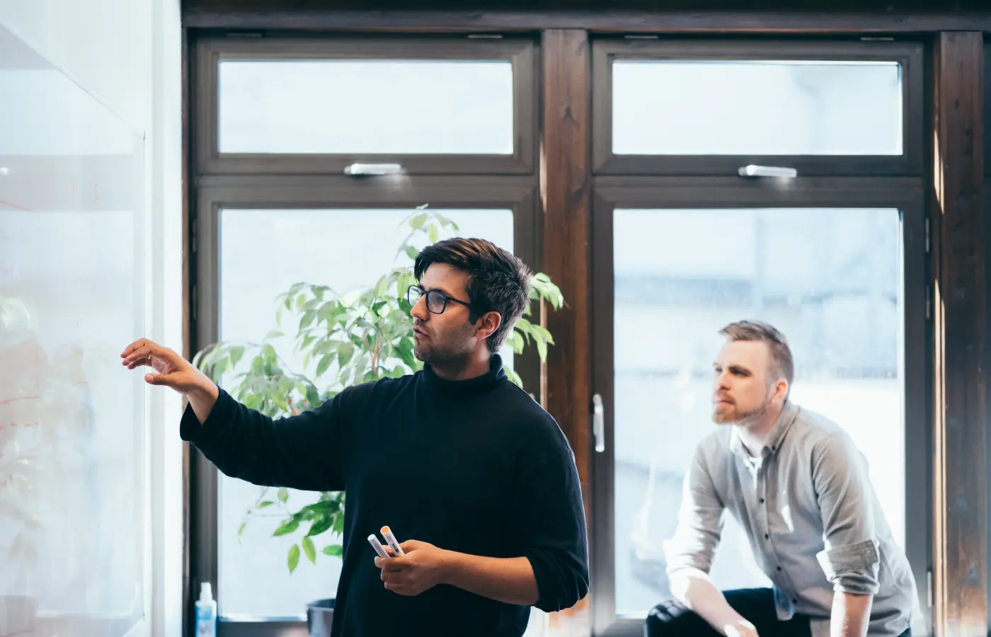 A man in glasses and a black sweater writing on a whiteboard while another man in a gray shirt attentively watches, with large windows and a green plant in the background.