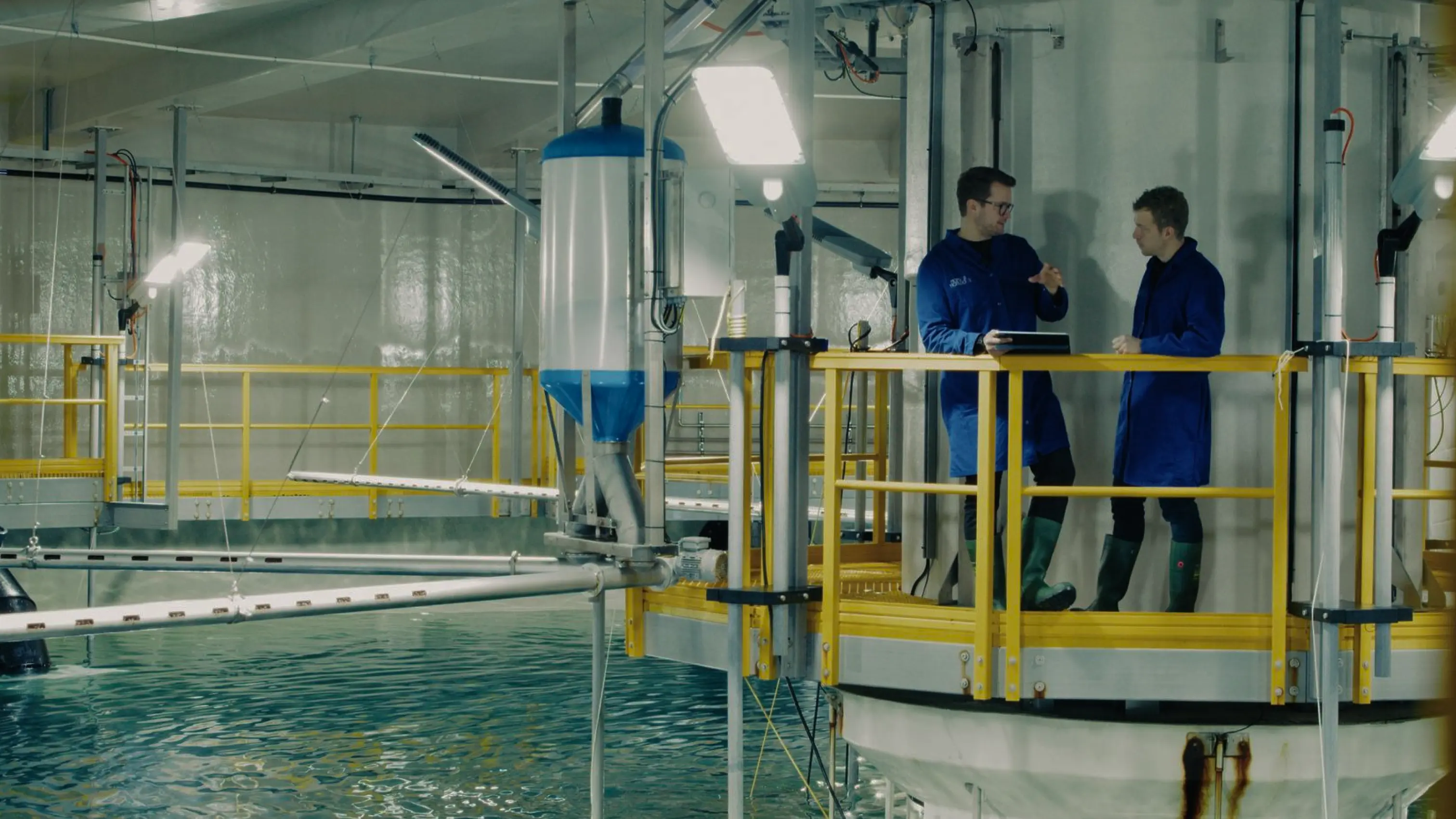 Two men wearing blue lab coats and green rubber boots standing on a yellow-railed platform above a large water tank, discussing work inside an industrial facility.