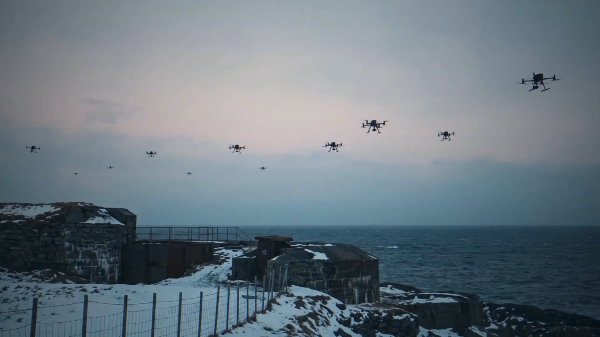 A fleet of drones flying in formation over a snowy, rocky coastal fortress with the ocean in the background under a cloudy sky.