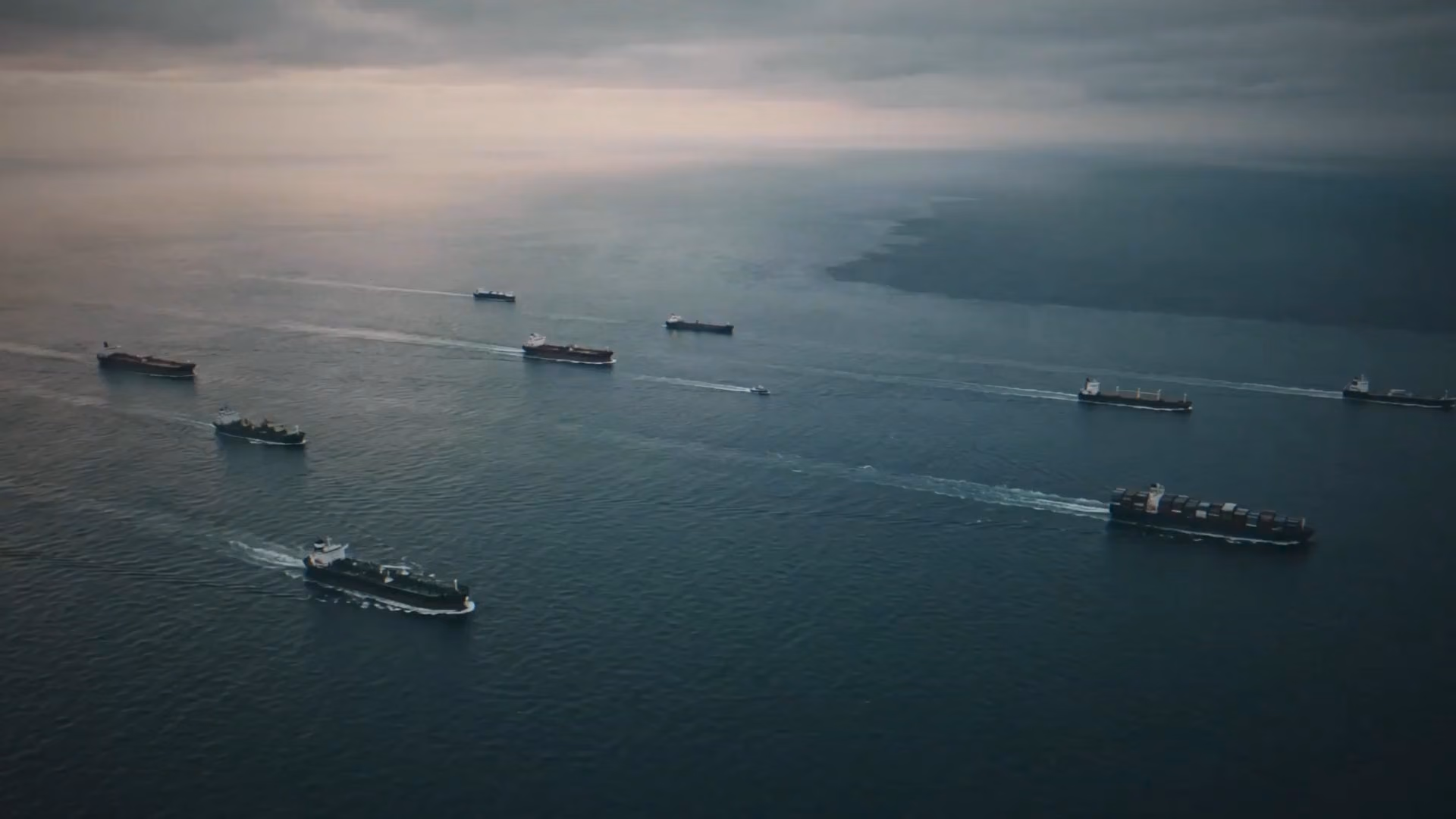 Aerial view of multiple cargo ships and boats navigating calm ocean waters under a cloudy sky during dusk or dawn.