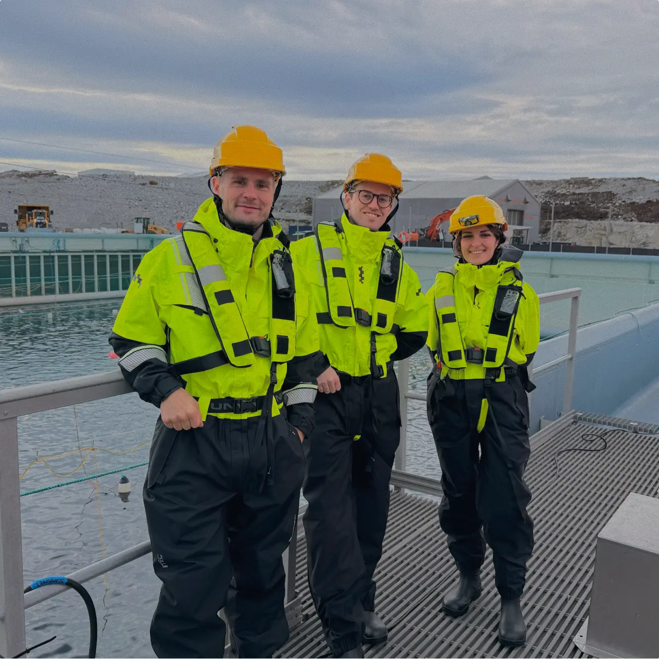 Three engineers in bright yellow safety jackets, black waterproof pants, and yellow helmets standing on a metal platform by a large water tank with industrial buildings and equipment in the background under a cloudy sky.