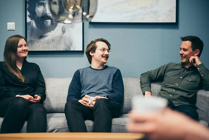 Three people sitting on a couch in a relaxed setting, smiling and engaged in conversation, with framed artwork on the wall behind them.