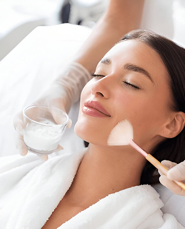 A woman with closed eyes receiving a facial treatment while a person wearing white gloves applies cream to her cheek with a brush.