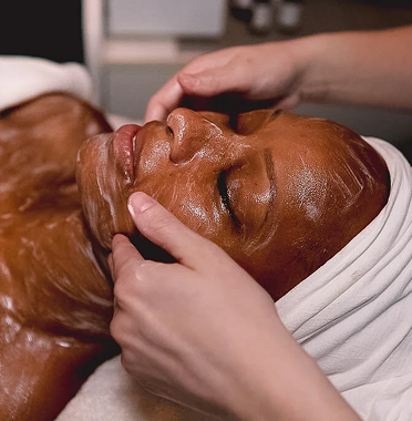 Person receiving a chocolate-colored facial treatment while lying down with a white towel wrapped around their head, and another person gently applying the treatment on their face using their hands.