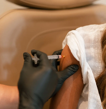 Person receiving an injection in the upper arm, administered by a healthcare worker wearing black gloves.