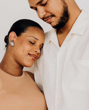 A woman with closed eyes and a slight smile resting her head on a man's chest, who is looking down at her with a gentle expression, both wearing light-colored tops.