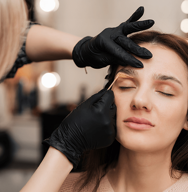 Close-up of a woman with closed eyes receiving eyebrow grooming by a person wearing black gloves using a small tool on her eyebrows in a softly lit indoor setting.