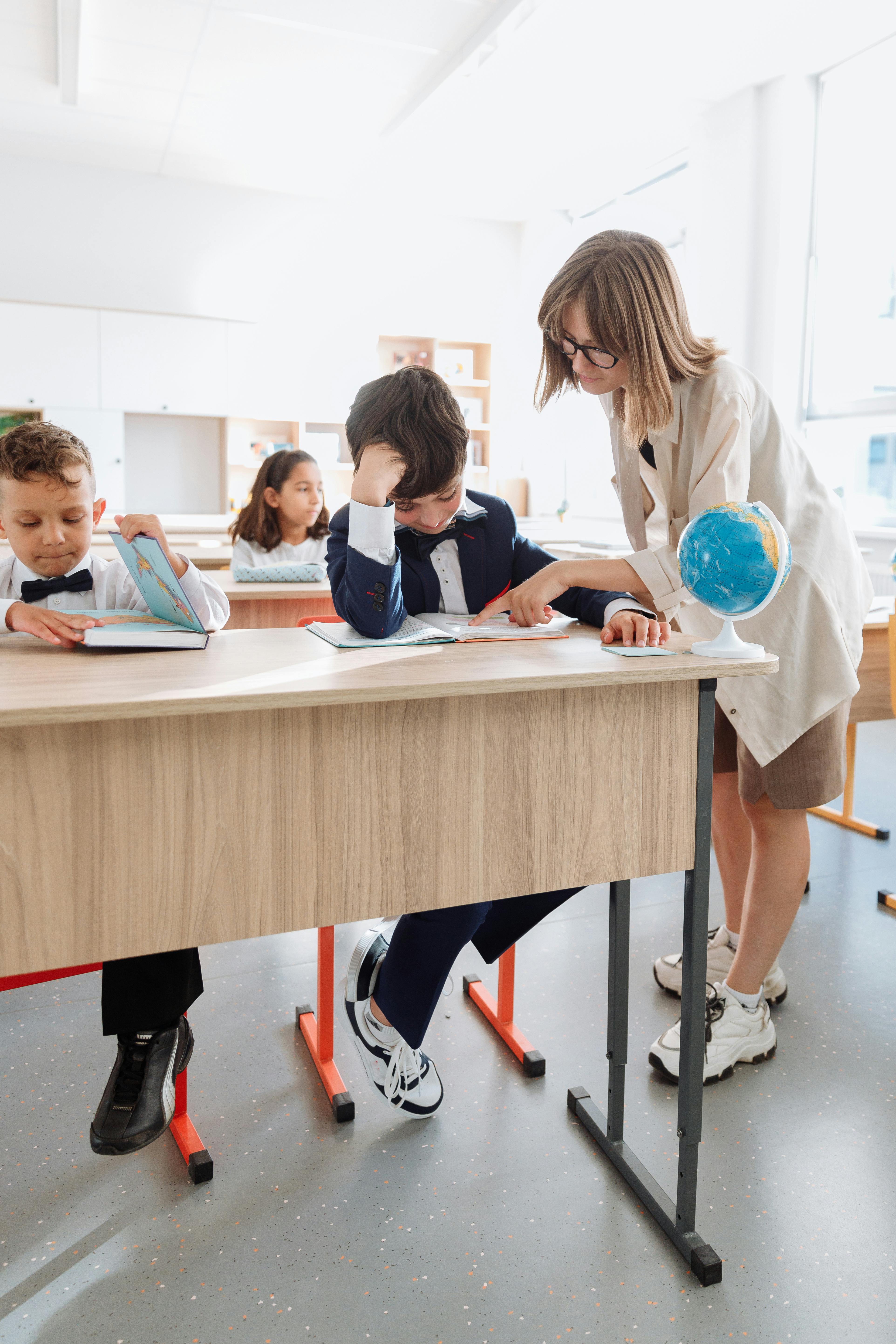 Teacher with glasses leaning over a student at a classroom desk, pointing at a book while two other children sit nearby, one reading and the other looking forward, with a globe on the table.
