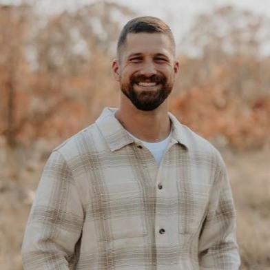 Smiling man with short brown hair and beard, wearing a beige plaid shirt, standing outdoors with blurred autumn trees in the background.