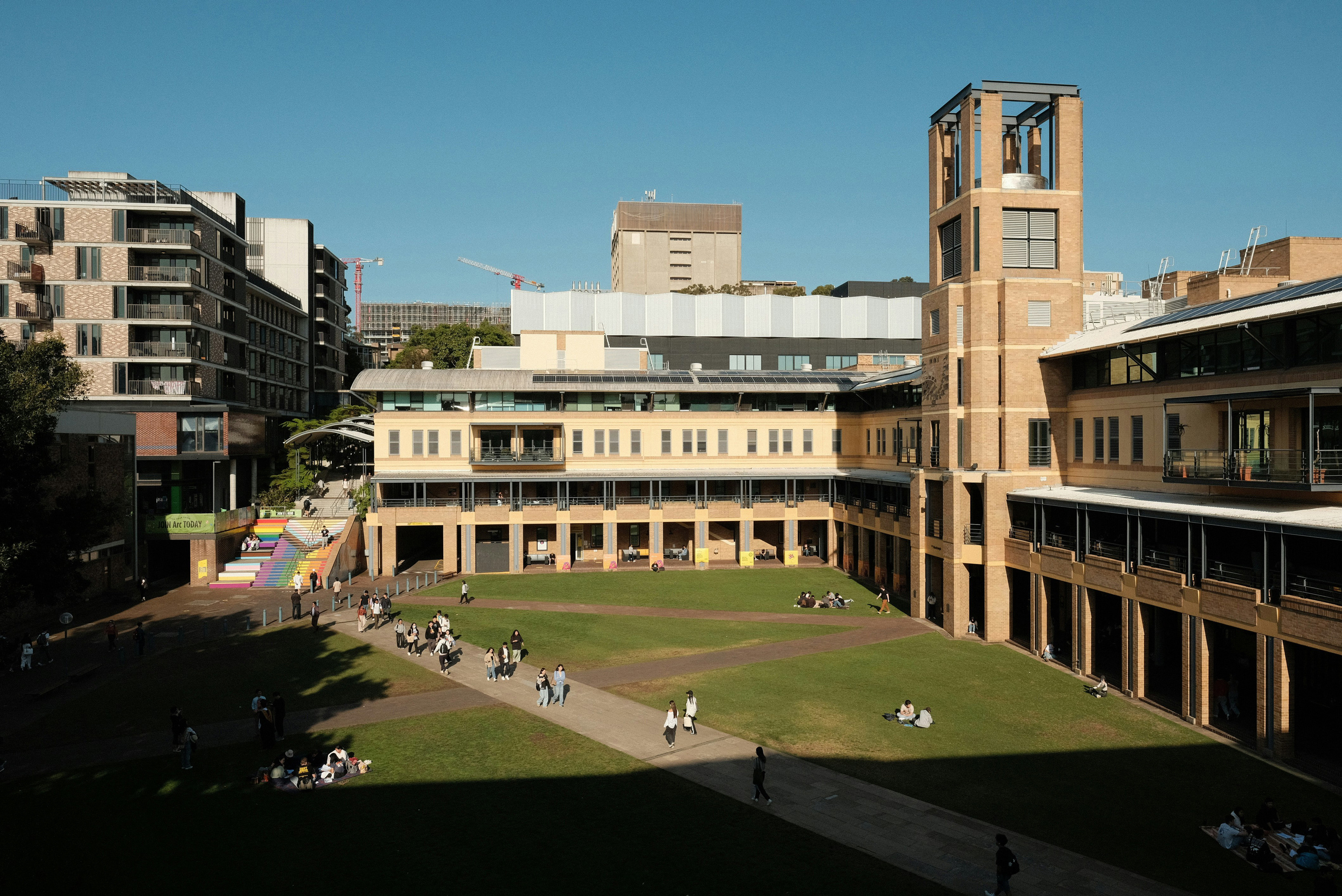 A university campus courtyard with students walking on paved paths and sitting on grass, surrounded by multi-story academic buildings under a clear blue sky.
