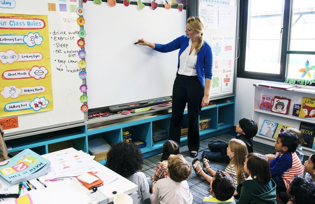 Teacher in blue cardigan writing on a whiteboard while a group of young students sit on the floor watching attentively in a brightly decorated classroom with posters and books.